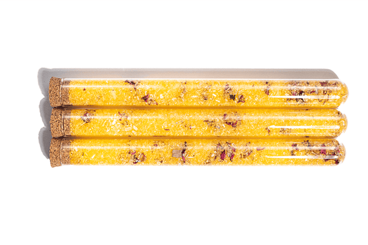 Image of 3 yellow bath salts in clear glass tubes with Rose Petals. The salts appear to have a fine, powdery texture and are a vibrant shade of yellow. Dried rose petals are visible inside the tubes, adding a decorative touch to the product. The tubes are arranged in a neat row against a white background.