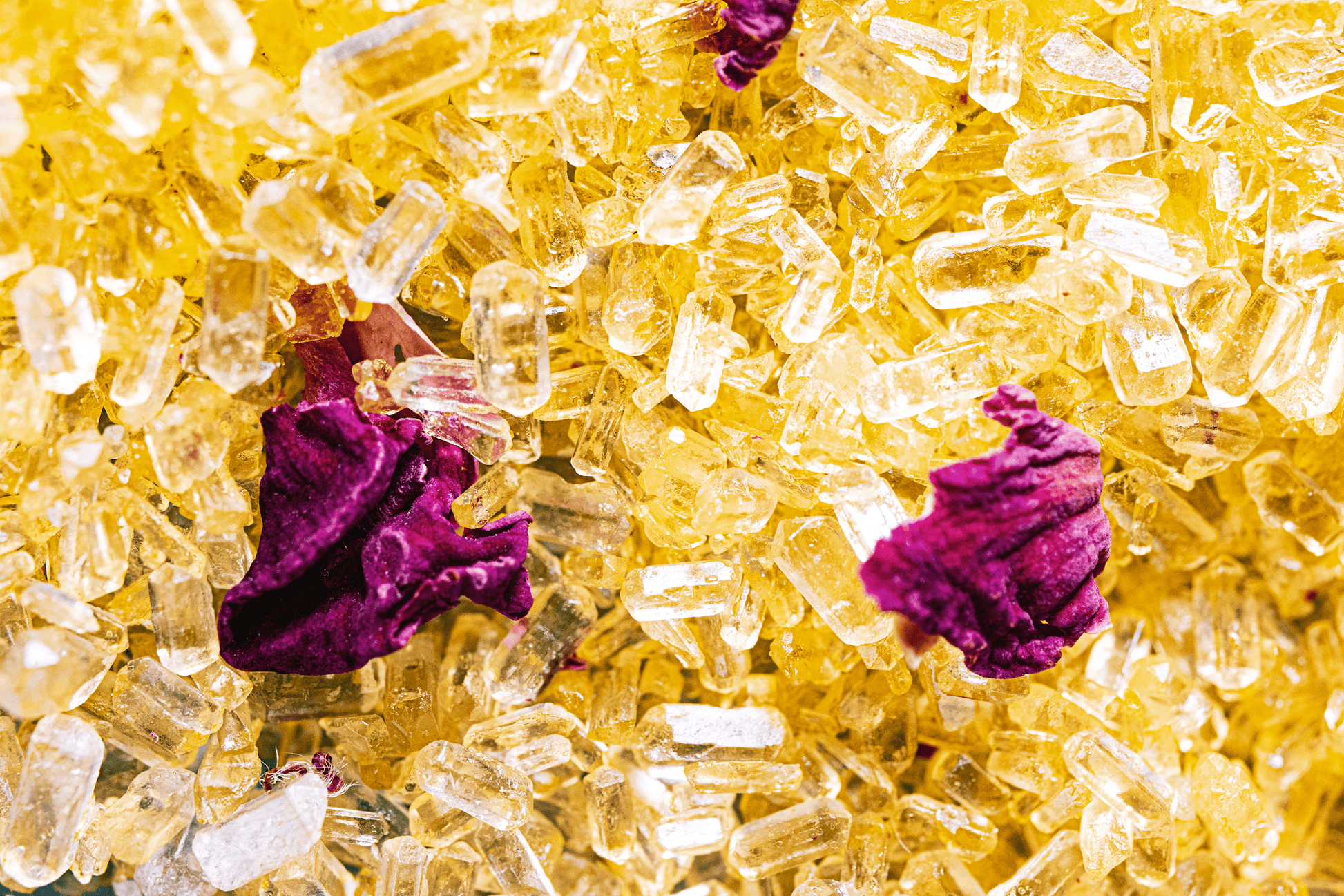 Close-up image of yellow bath salts with dried rose petals on top, arranged in a small pile. The salts have a fine, powdery texture and the yellow color is bright and sunny. The rose petals are mostly pink with some red accents and add a decorative element to the salts. The background is blurred and out of focus.