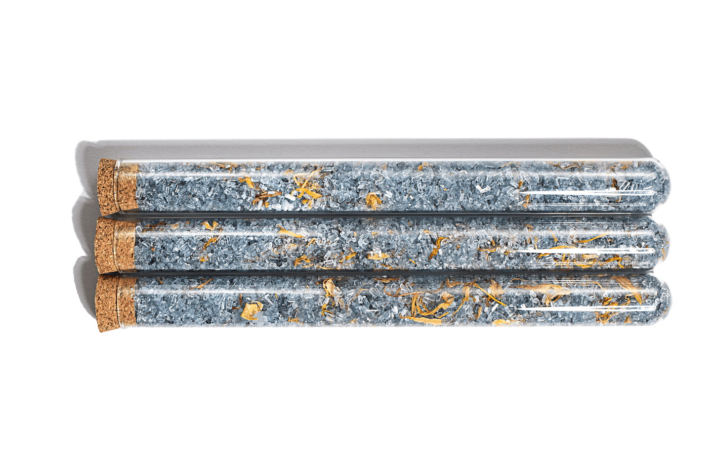 Image of 3 grey bath salts in clear glass tubes with Calendula. The salts appear to have a fine, powdery texture and are a subtle shade of grey. Dried calendula flowers are visible inside the tubes, adding a pop of color and decorative touch to the product. The tubes are arranged in a neat row against a white background