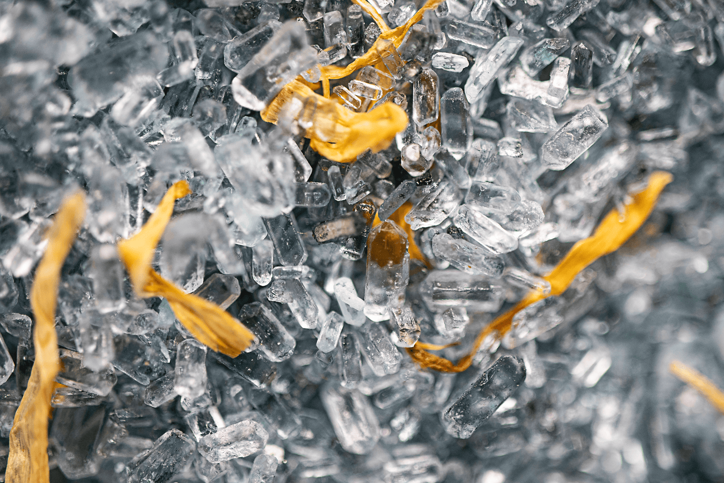 Close-up image of grey bath salts with dried calendula flowers on top, arranged in a small pile. The salts have a fine, powdery texture and the color is a soft shade of grey. The calendula flowers are a bright orange-yellow color and add a decorative element to the salts. The background is blurred and out of focus