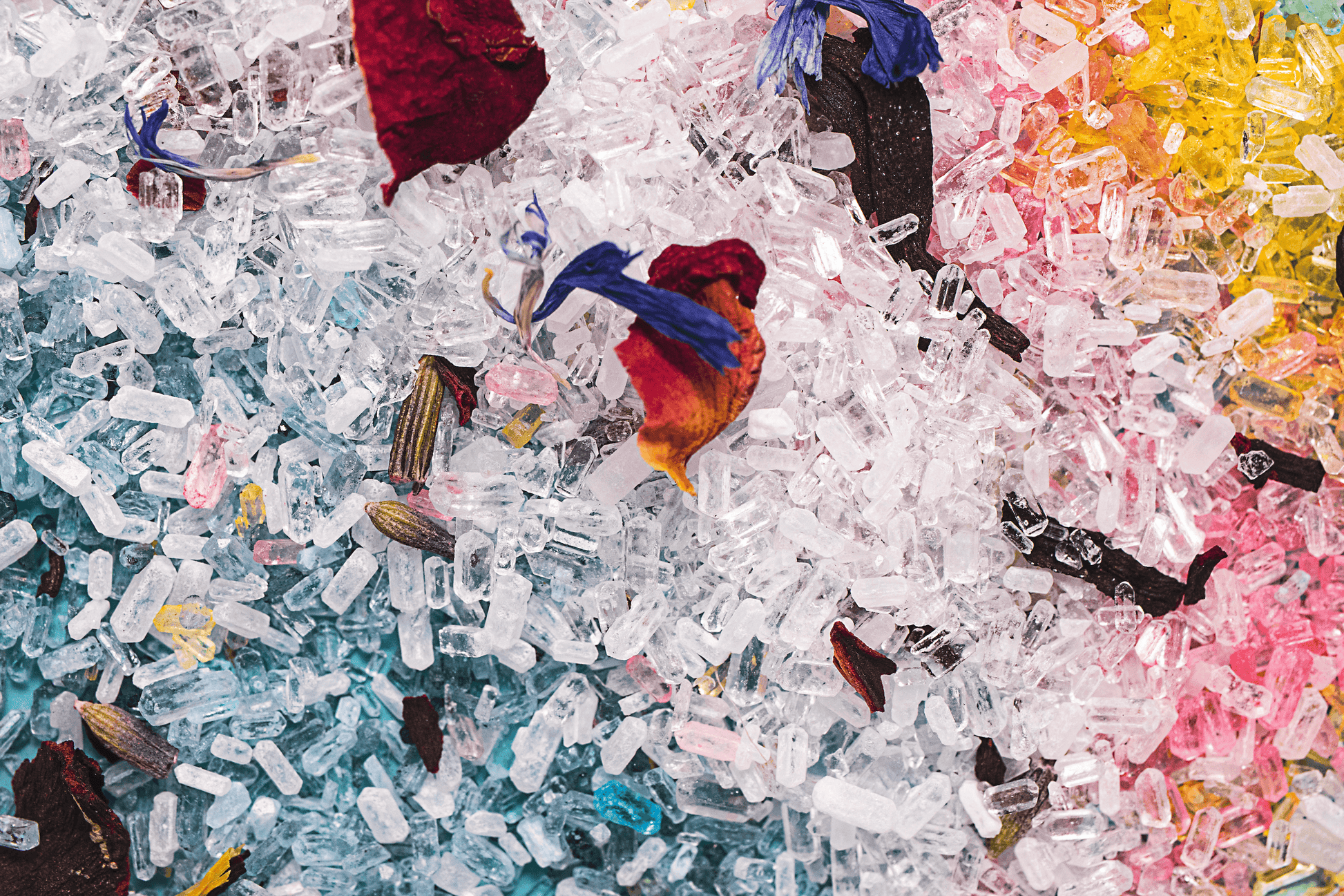 Close-up image of a mix of colorful bath salts with various dried flower petals on top, arranged in a small pile. The salts have a fine, powdery texture and come in shades of pink, blue, yellow, white, and pink. Dried petals from flowers such as roses, cornflowers, hibiscus, lavender, and calendula are visible on top of the salts, adding a decorative element to the product. The background is blurred and out of focus