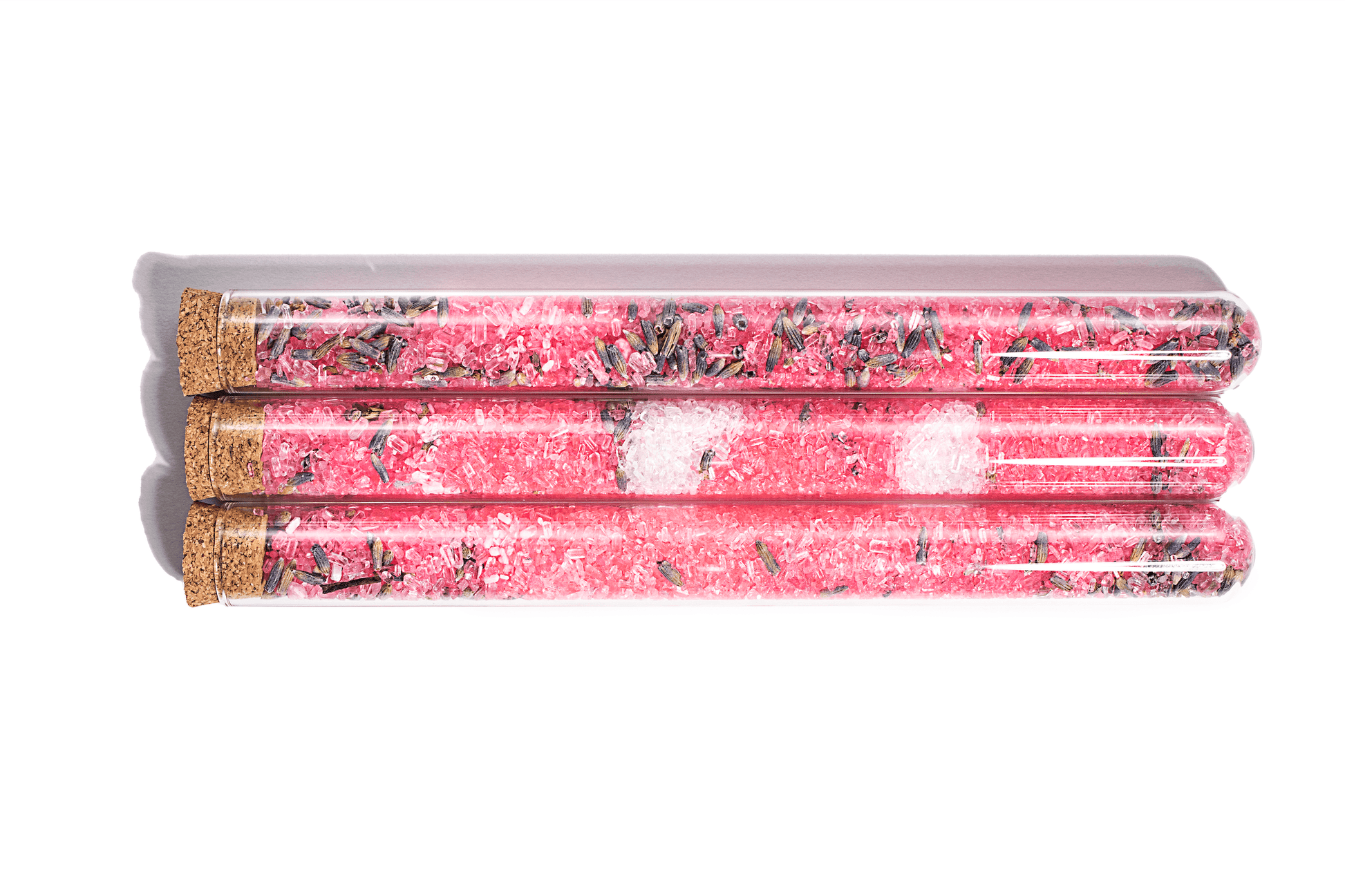 Image of pink bath salts in clear glass tubes with Lavender. The salts appear to have a fine, powdery texture and are a delicate shade of pink. Dried lavender buds are visible inside the tubes, adding a touch of purple and a pleasant aroma to the product. The tubes are arranged in a neat row against a white background