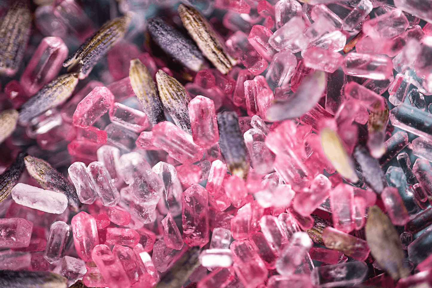 Close-up image of pink bath salts with dried lavender buds on top, arranged in a small pile. The salts have a fine, powdery texture and the color is a soft shade of pink. The lavender buds are a deep purple color and add a decorative element to the salts, as well as a pleasant aroma. The background is blurred and out of focus